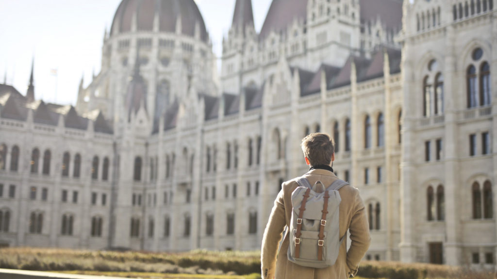 male wearing brown coat backpack near hungarian parliament building budapest hungary copy